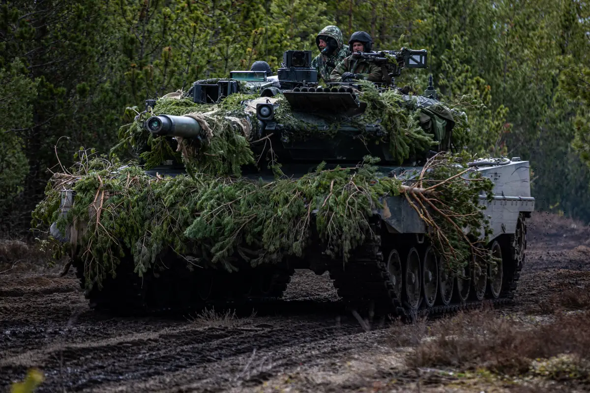 A camouflaged Leopard 2 tank on the move along a forest track, with its crew in the turret.