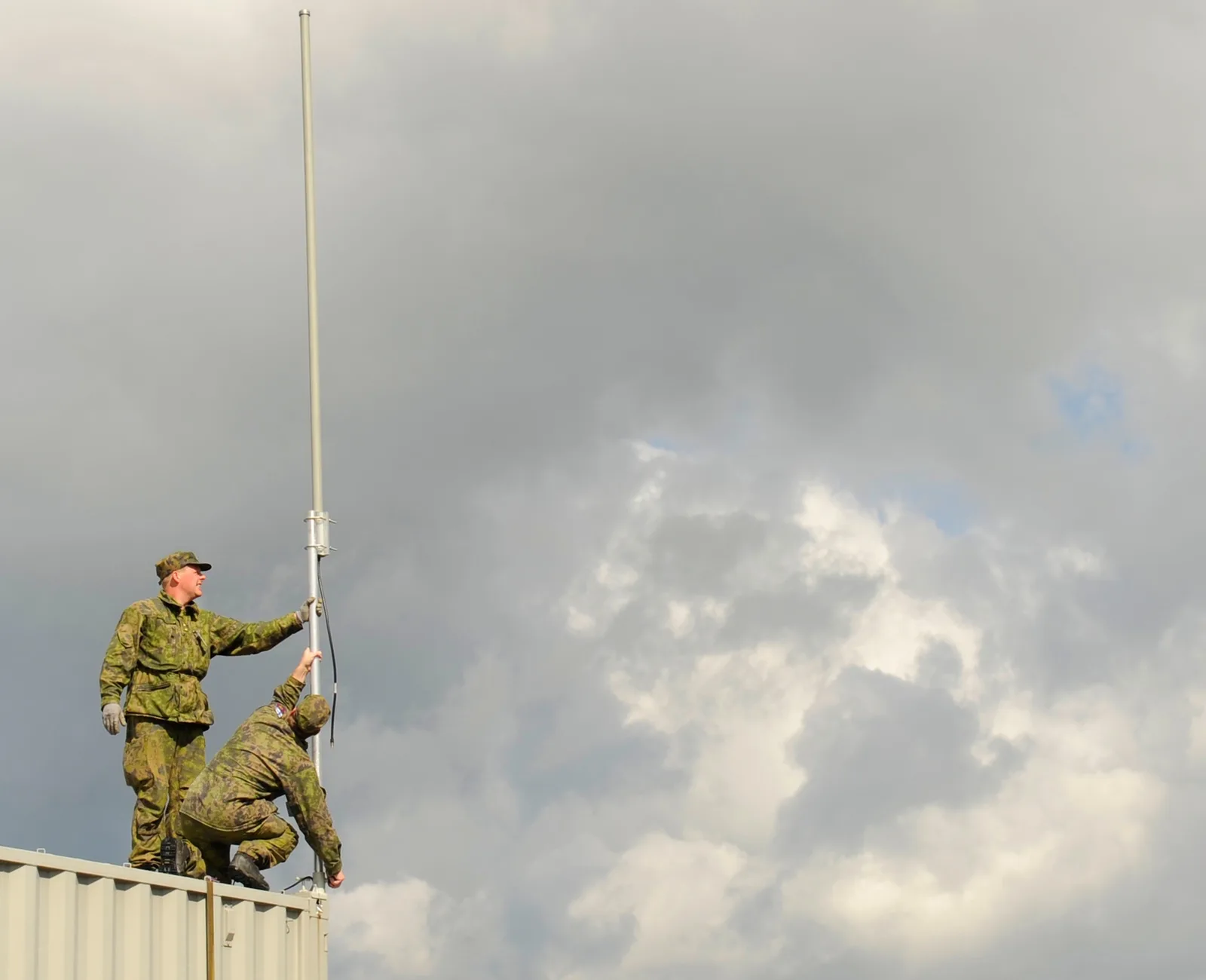 Two soldiers raising a whip antenna mast on the roof of a field shelter under storm clouds.